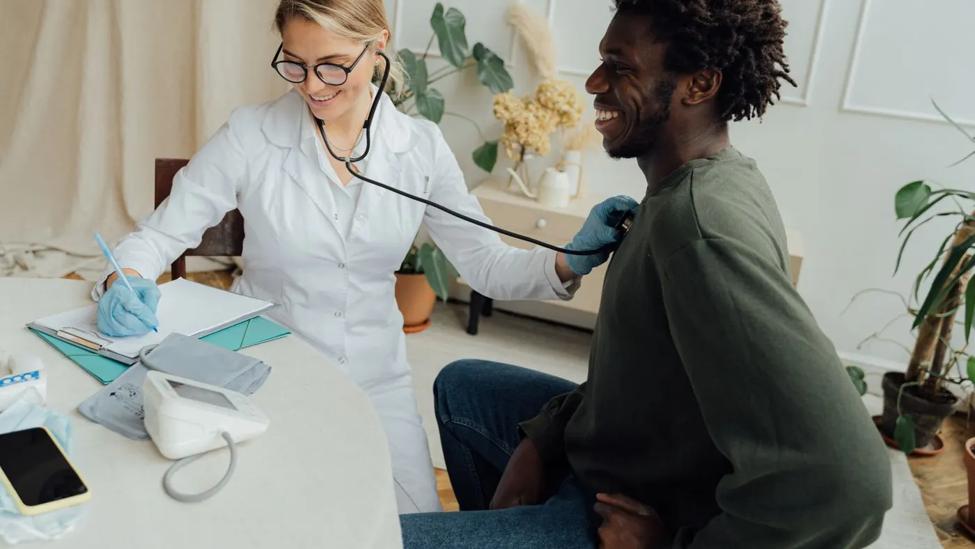 Médica con bata blanca y estetoscopio que revisa a un paciente sentado frente a una mesa con tensiómetro y carpeta: imagen para la guía del colesterol alto