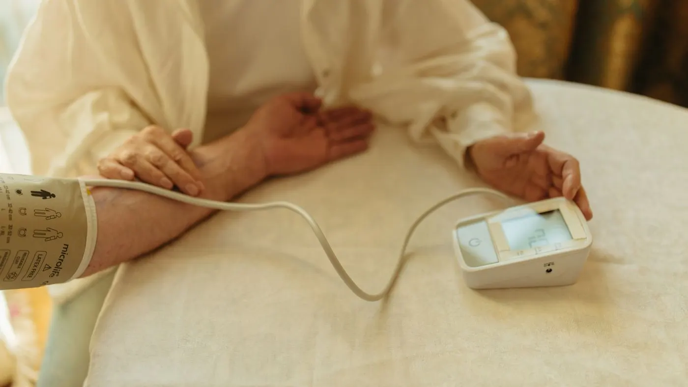 Antebrazo de una persona sentada a una mesa con el brazalete de un tensiómetro digital puesto y el aparato al lado: medición de la presión en casa