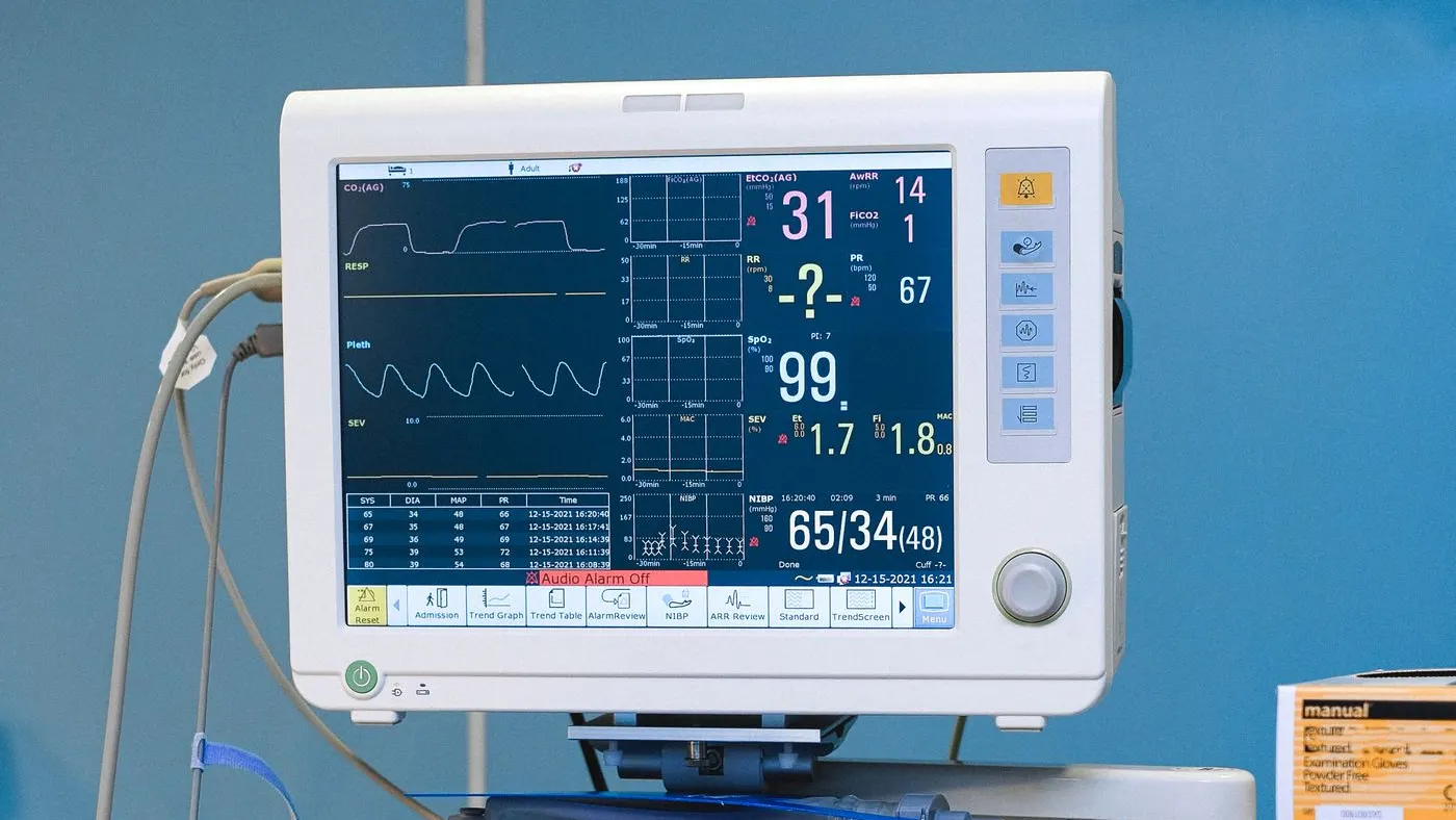 Monitor de signos vitales con pantalla oscura, trazos y cifras de colores, frente a una pared azul de un quirófano: imagen para la guía de los síntomas de infarto