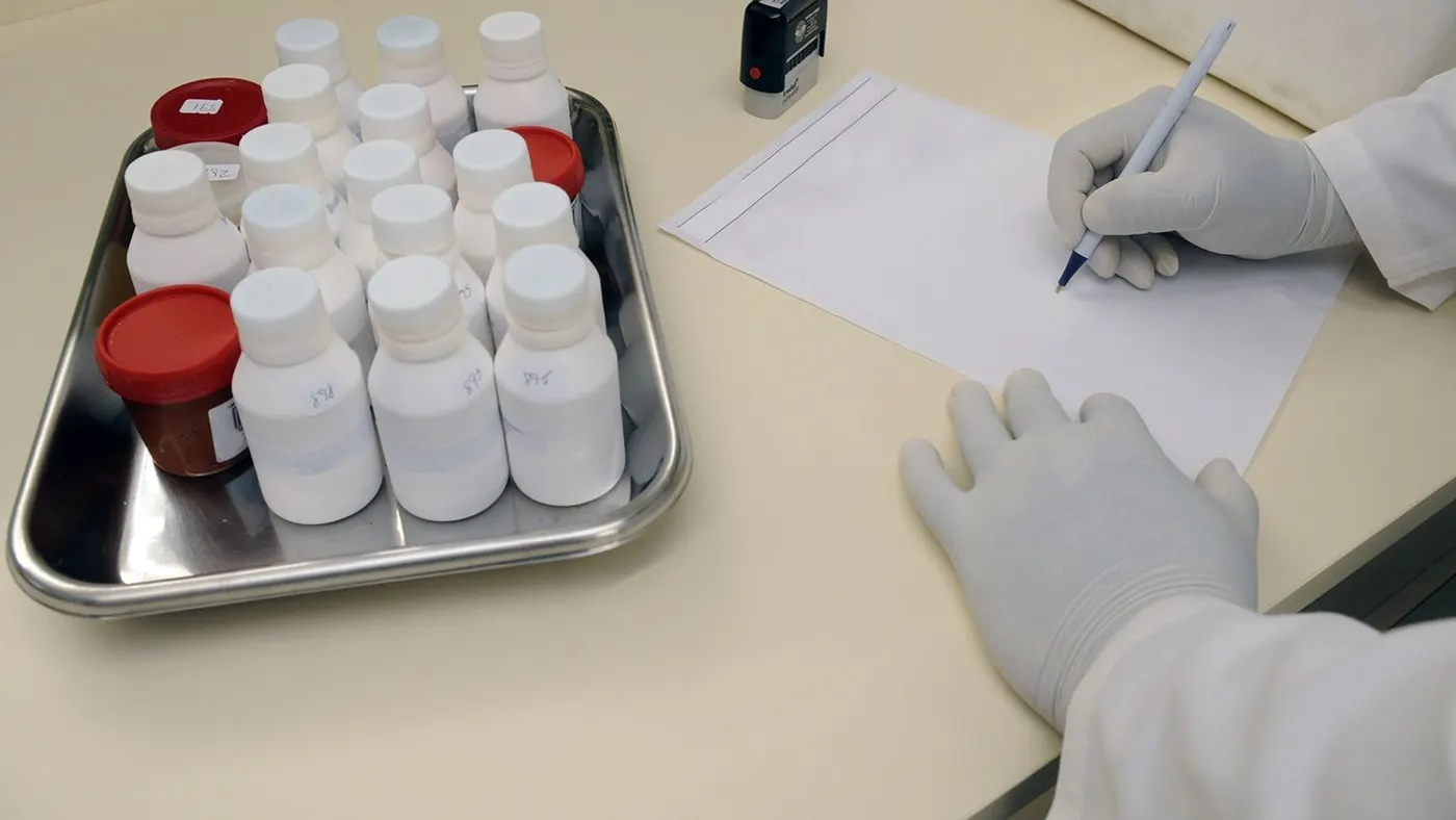 Bandeja metálica con frascos de muestras en un laboratorio y una mano con guante que anota en una hoja: imagen para la guía de los triglicéridos